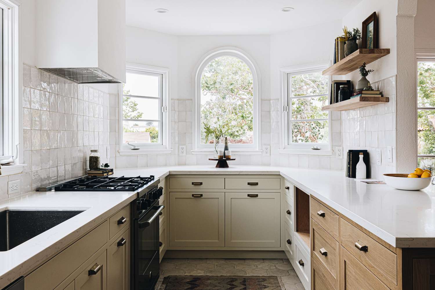 Bright modern kitchen with shaker-style lower cabinets, white countertops, floating wood shelves, and an arched window, showcasing a premium kitchen cabinet supplier design.