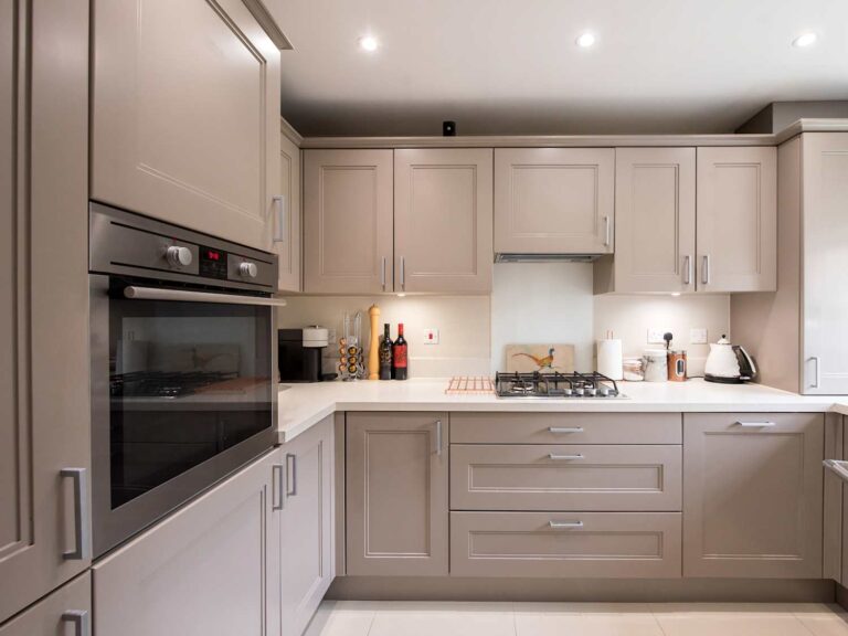 Modern beige shaker kitchen cabinets in an L-shaped layout with built-in oven, gas cooktop, and white countertops.