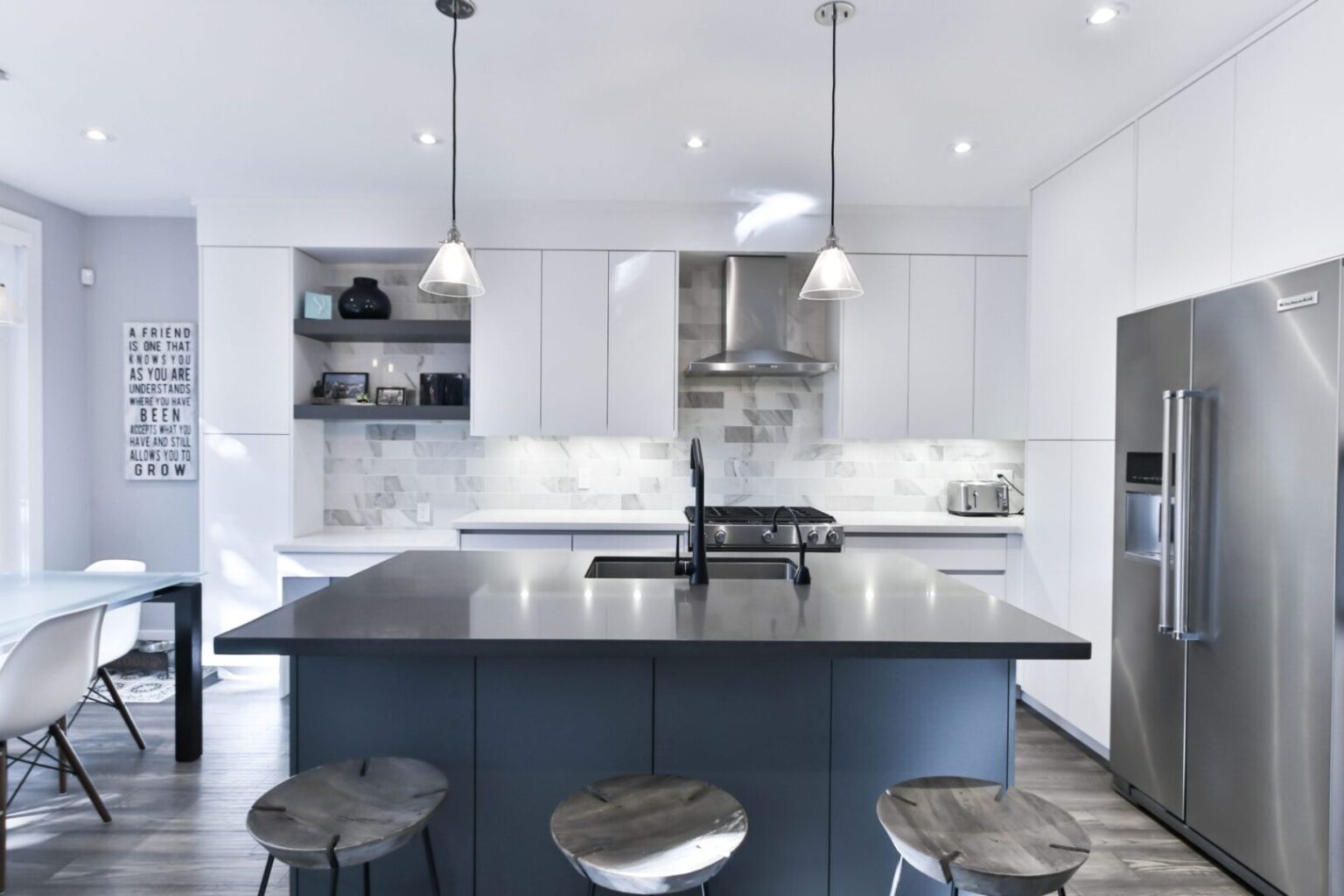 Modern white flat-panel kitchen cabinets with a large dark island, pendant lights, and stainless-steel appliances, supplied by a trusted kitchen cabinet supplier.