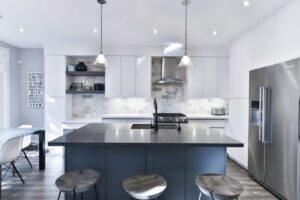 Modern white flat-panel kitchen cabinets with a large dark island, pendant lights, and stainless-steel appliances, supplied by a trusted kitchen cabinet supplier.
