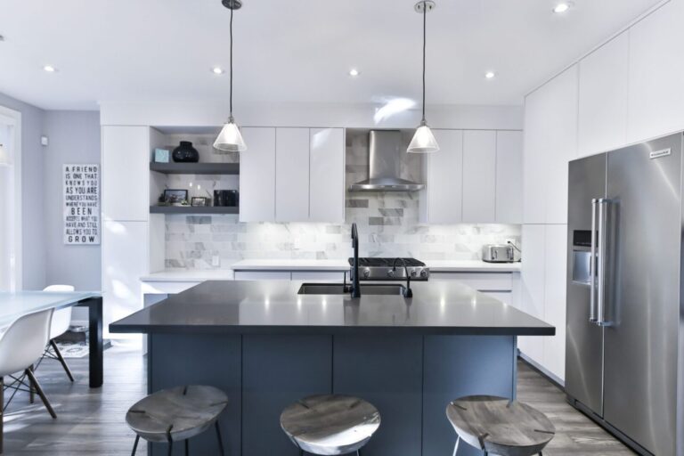 Modern white flat-panel kitchen cabinets with a large dark island, pendant lights, and stainless-steel appliances, supplied by a trusted kitchen cabinet supplier.