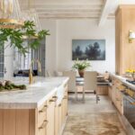 Sunlit kitchen cabinets in natural wood with brass hardware, marble countertops, and a large modern island