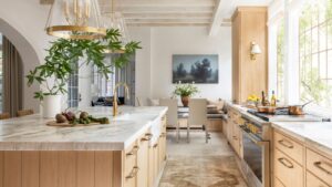 Sunlit kitchen cabinets in natural wood with brass hardware, marble countertops, and a large modern island