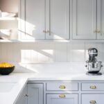 Kitchen Cabinets Hamilton in a bright modern kitchen with light gray shaker cabinets, brass hardware, white countertops, and natural sunlight.