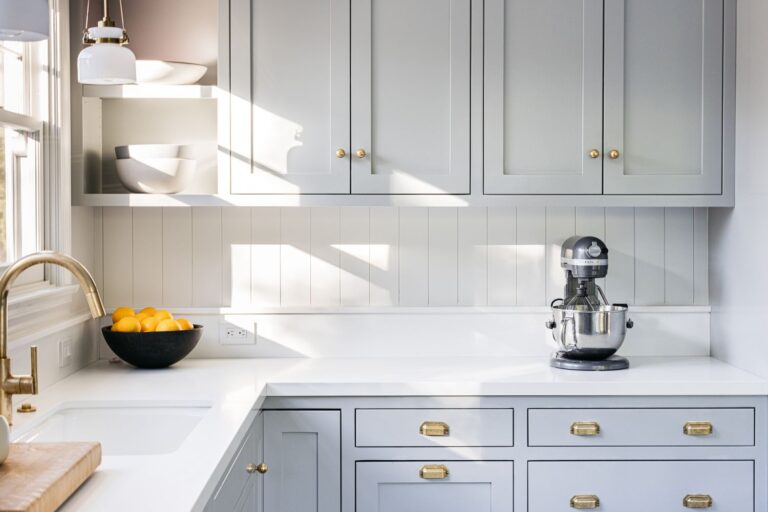 Kitchen Cabinets Hamilton in a bright modern kitchen with light gray shaker cabinets, brass hardware, white countertops, and natural sunlight.