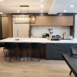 Modern Kitchen Cabinets Etobicoke featuring a large white waterfall island, black lower cabinets, light wood upper cabinetry, and elegant pendant lighting in an open-concept home.