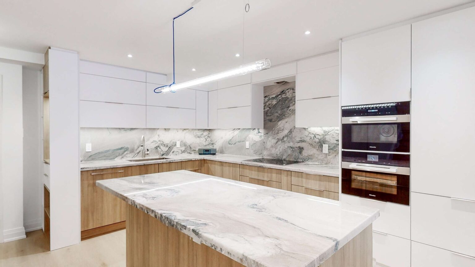 Contemporary Kitchen Cabinets Toronto design with sleek white upper cabinets, warm wood lower cabinets, marble-look countertops, a waterfall island, and built-in wall ovens in a bright modern space.