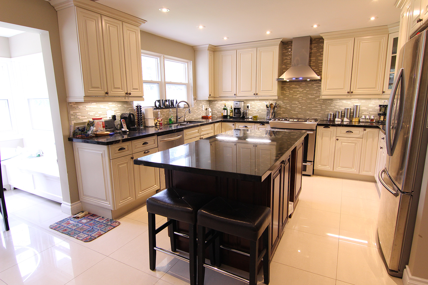 Modern kitchen cabinets Mississauga home featuring cream raised-panel cabinetry, a large dark island, stainless steel appliances, and a tile backsplash.