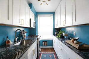 White shaker kitchen cabinets Mississauga in a bright galley kitchen with brass handles, blue walls, and dark marble countertops