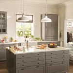 Bright farmhouse-style kitchen with gray shaker IKEA kitchen cabinets, a large drawer island, white countertops, pendant lights, and white subway-tile walls.