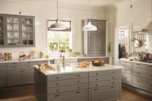 Bright farmhouse-style kitchen with gray shaker IKEA kitchen cabinets, a large drawer island, white countertops, pendant lights, and white subway-tile walls.
