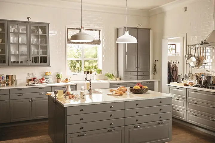 Bright farmhouse-style kitchen with gray shaker IKEA kitchen cabinets, a large drawer island, white countertops, pendant lights, and white subway-tile walls.