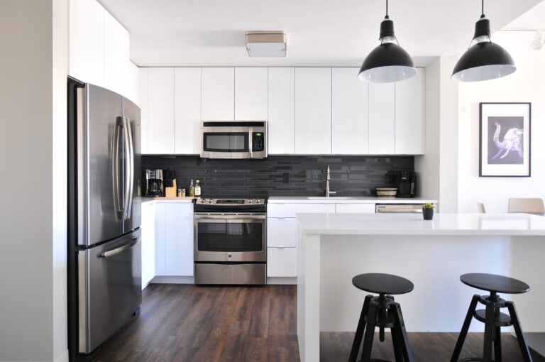 Minimalist white kitchen with flat-panel cabinets, black backsplash, stainless steel appliances, and pendant lights, illustrating a sleek contemporary layout for what are IKEA kitchen cabinets made of content.