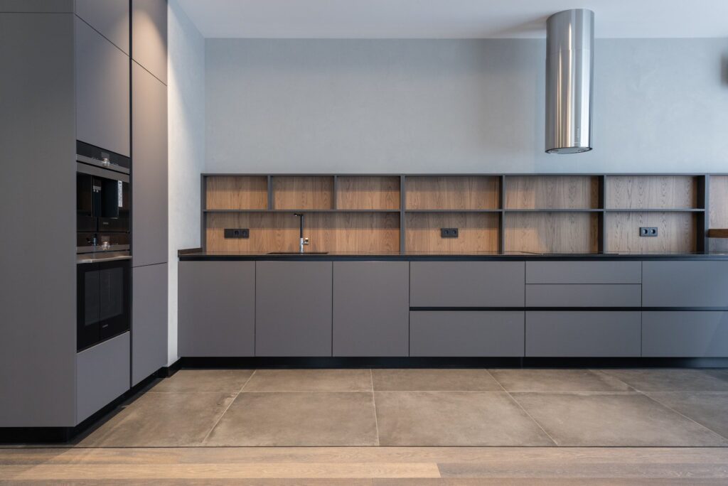 Modern minimalist kitchen featuring IKEA style kitchen cabinets in matte gray with flat-panel doors, black countertop, wood backsplash, and built-in appliances.