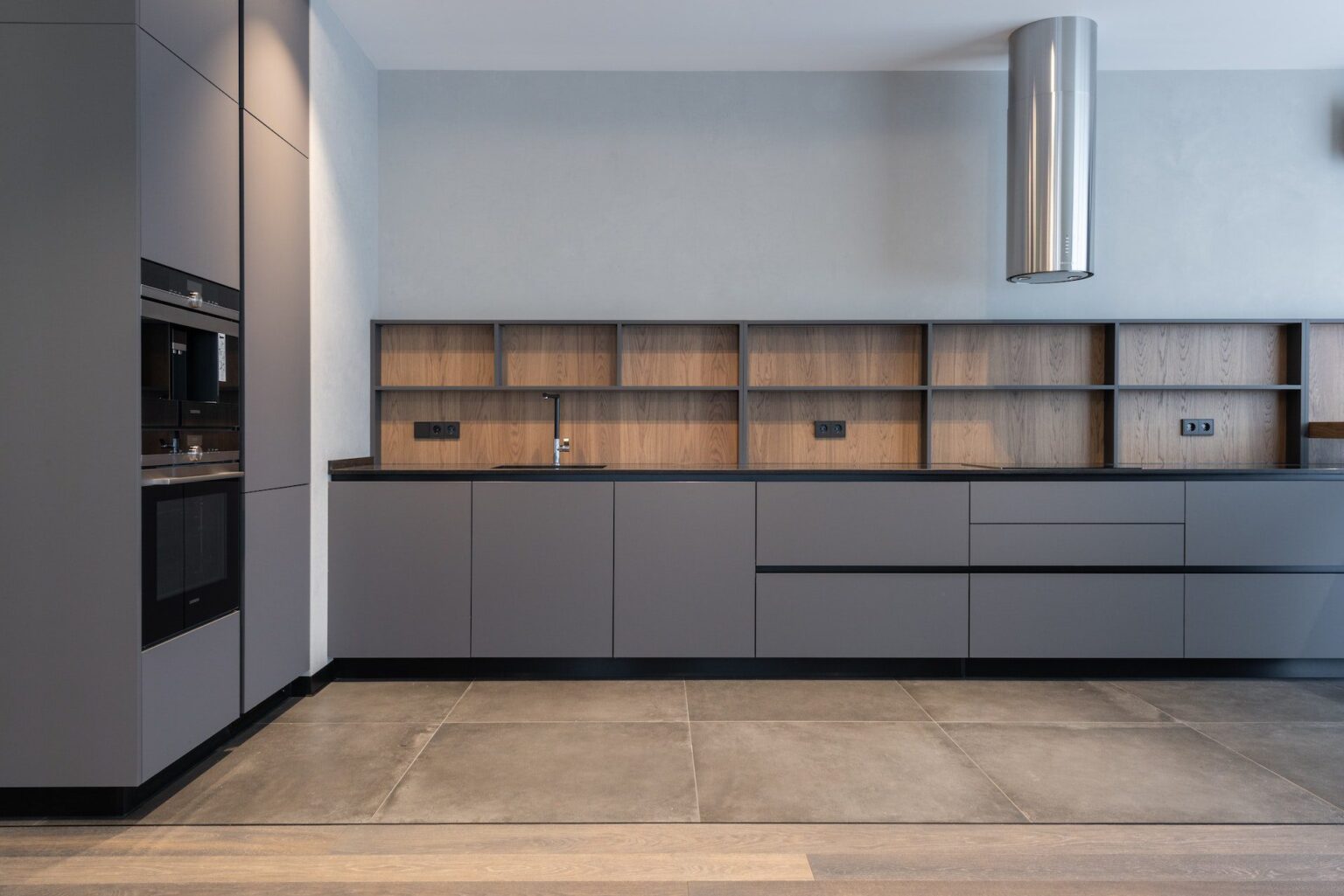 Modern minimalist kitchen featuring IKEA style kitchen cabinets in matte gray with flat-panel doors, black countertop, wood backsplash, and built-in appliances.